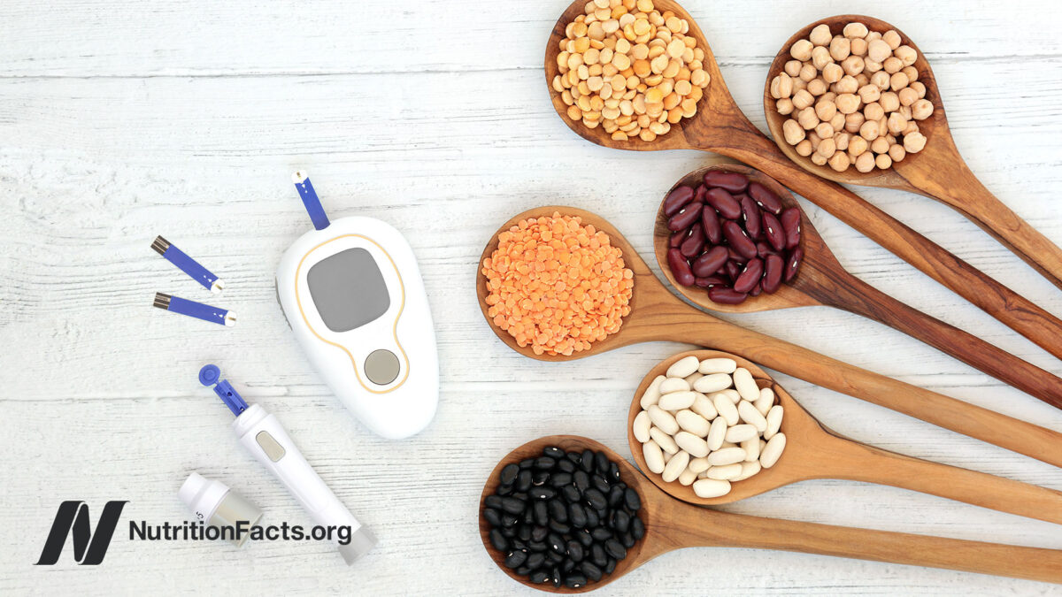 Two blood glucose meters surrounded by a few testing strips next to wooden serving spoons full of various beans and legumes