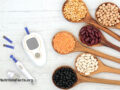 Two blood glucose meters surrounded by a few testing strips next to wooden serving spoons full of various beans and legumes