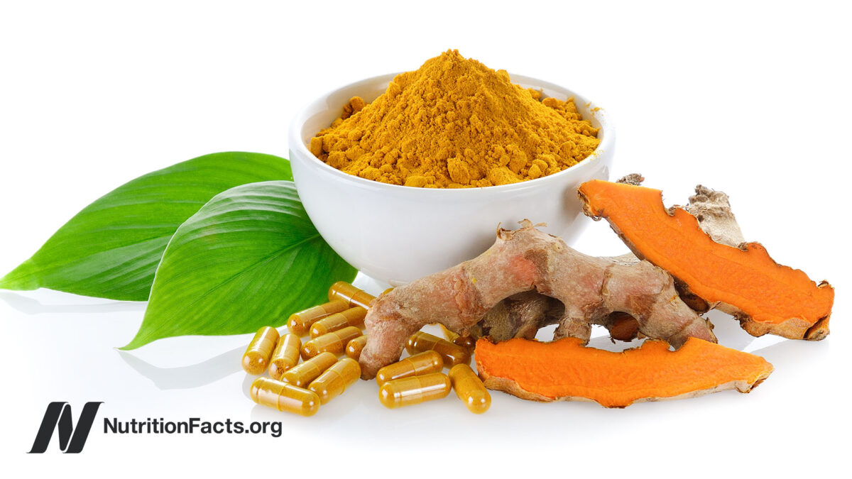 White bowl of ground turmeric powder behind fresh turmeric roots and turmeric supplement capsules