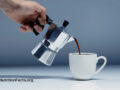 Hand holding a Moka pot pouring hot coffee into a white mug