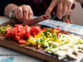 Elder hands using a knife to cut fresh vegetables like tomatoes and lettuce on a wooden cutting board
