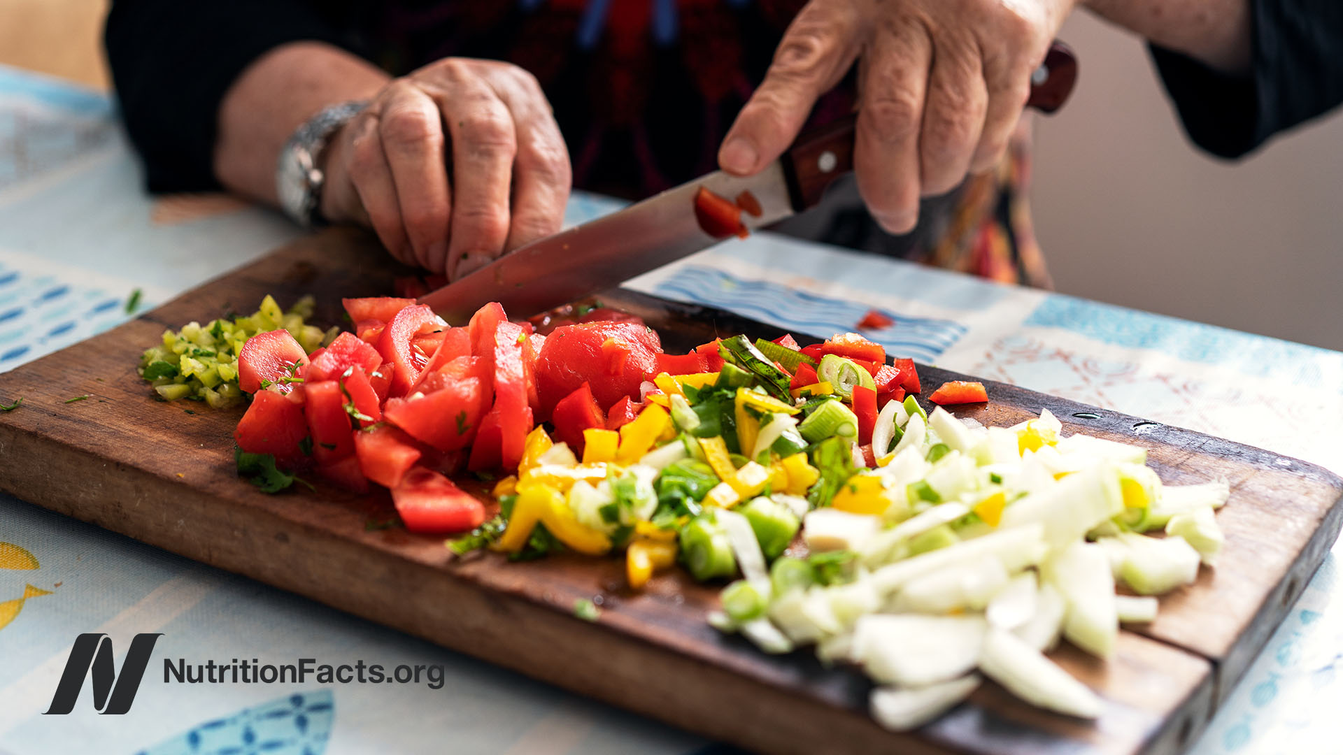 Elder hands using a knife to cut fresh vegetables like tomatoes and lettuce on a wooden cutting board