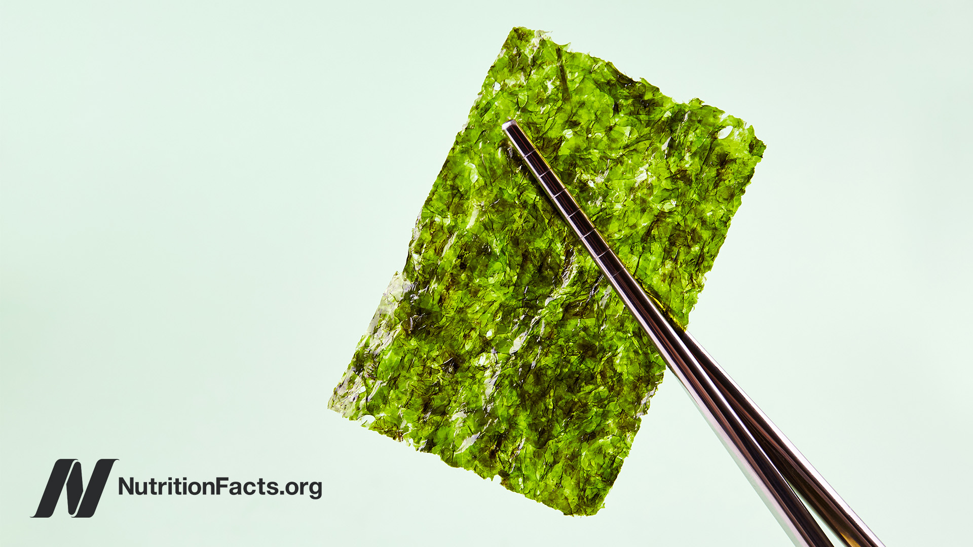 Dark wooden chopsticks holding up a dried seaweed nori sheet in front of a light