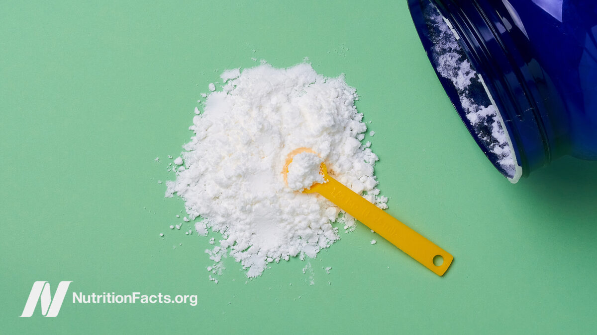 A large blue glass container on its side with spilled white taurine powder with a yellow measuring scoop in the center of the powder pile in front of a mint green background