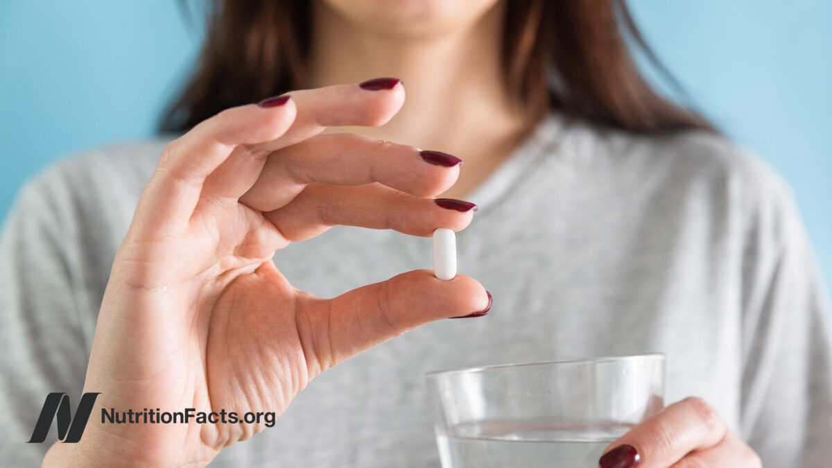 Hand with painted nails holding up a white statin tablet while the other hand is holding a glass of water