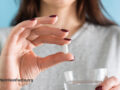 Hand with painted nails holding up a white statin tablet while the other hand is holding a glass of water