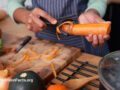 Person peeling a carrot in their kitchen surrounded by various vegetables